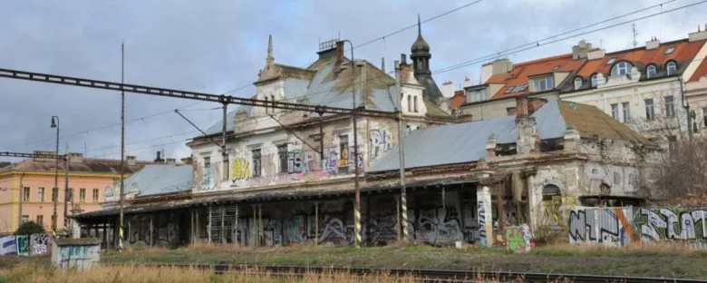 Zanedbaná budova nádraží s přilehlými kolejemi a zarostlou zemí.