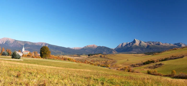Podzimní krajina s horami, poli a kostelem pod modrou oblohou.