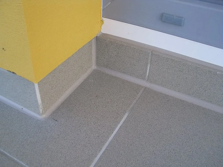 Detailní pohled na dlažbu v rohu balkonu s žlutou stěnou.